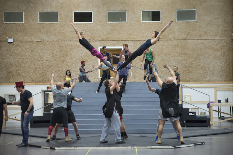 Acrobatics and performerson on blue mat in a rehearsal hall. Two woman held by their feet above their heads create a V shape.